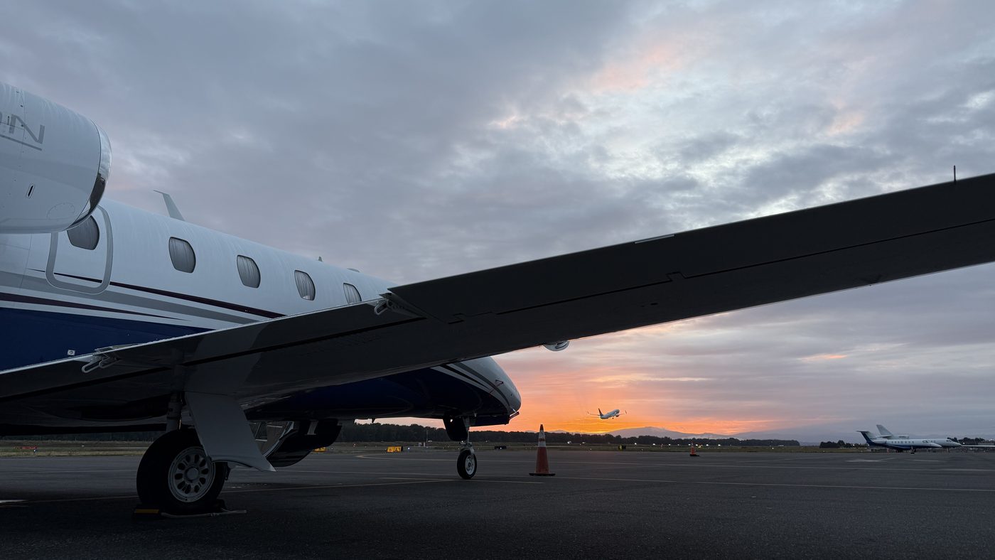 A Cessna Citation XLS+ parked on the ramp at sunrise, seen from under the wing, with an airliner departing in the distance
